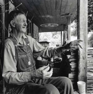 Ray telling a story on his front porch. Sticks nailed to the wall forecast the weather. The hex painted on the ceiling is to keep ghosts away which Rosa says, "works most of the time!" (Photo by: David Holt)